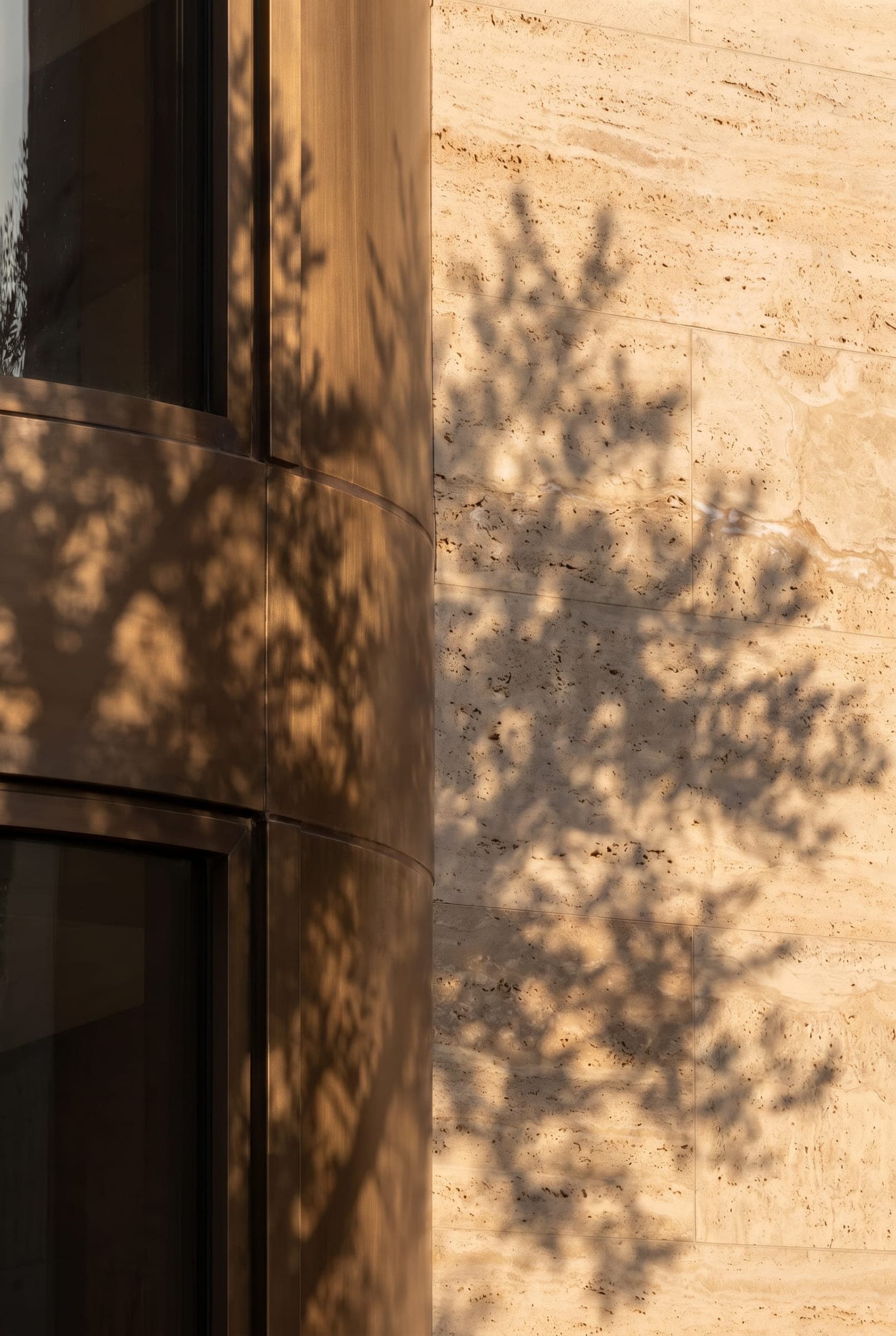 The shadow of a tree falling across a travertine wall beside a bronze-framed window in low sun — material study by LKYOOB