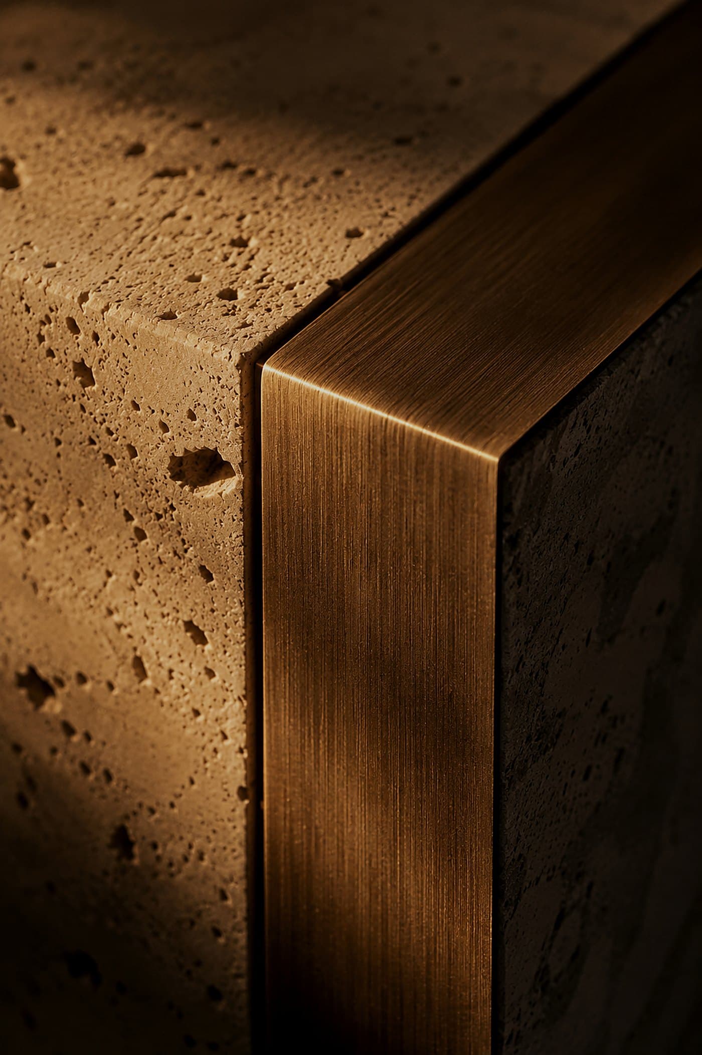 Brushed bronze edge meeting a travertine block at a precise junction under raking light — material study by LKYOOB