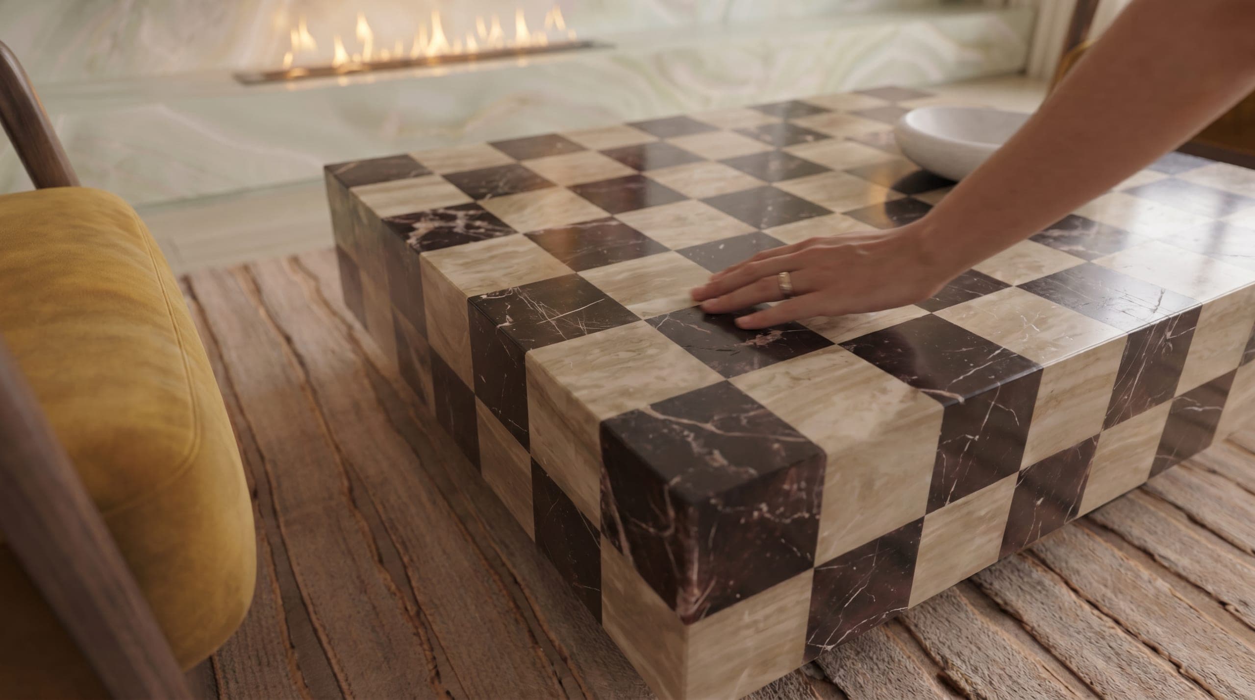 Macro of a book-matched Nero Marquina and travertine checkerboard table with a hand resting on it, fireplace glow beyond — bespoke craft by LKYOOB