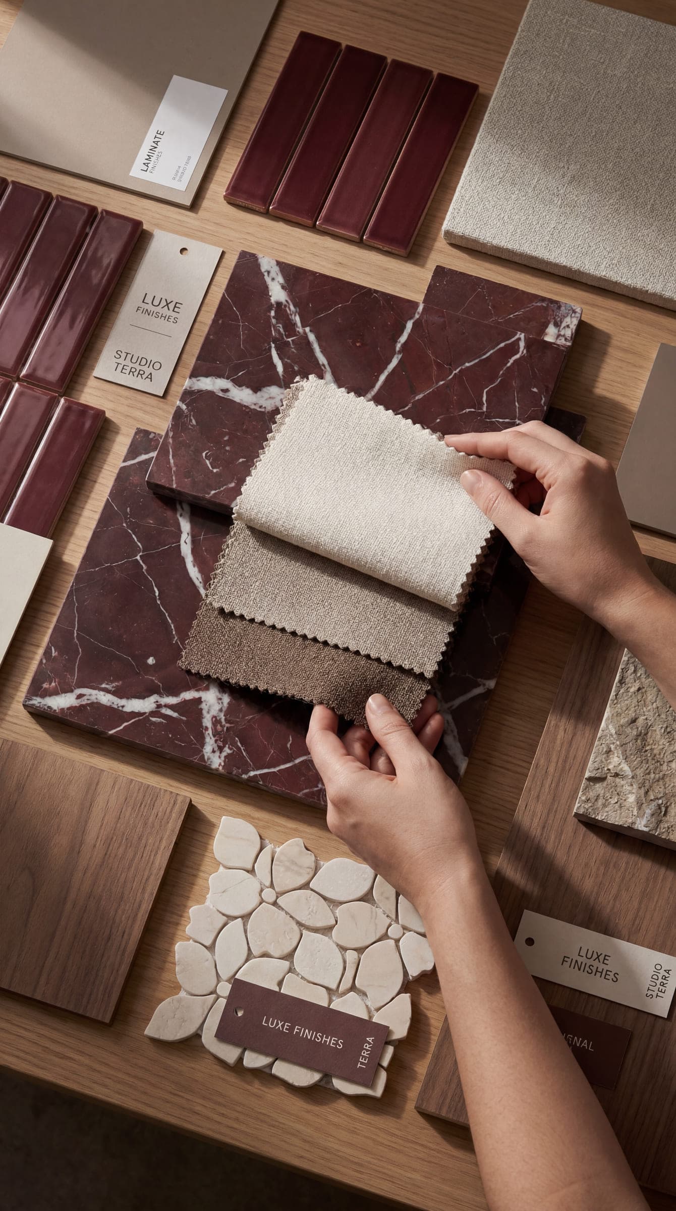 Stone, tile, timber and fabric samples laid out on a timber table, with three woven swatches held against a red marble slab — specifying materials at LKYOOB