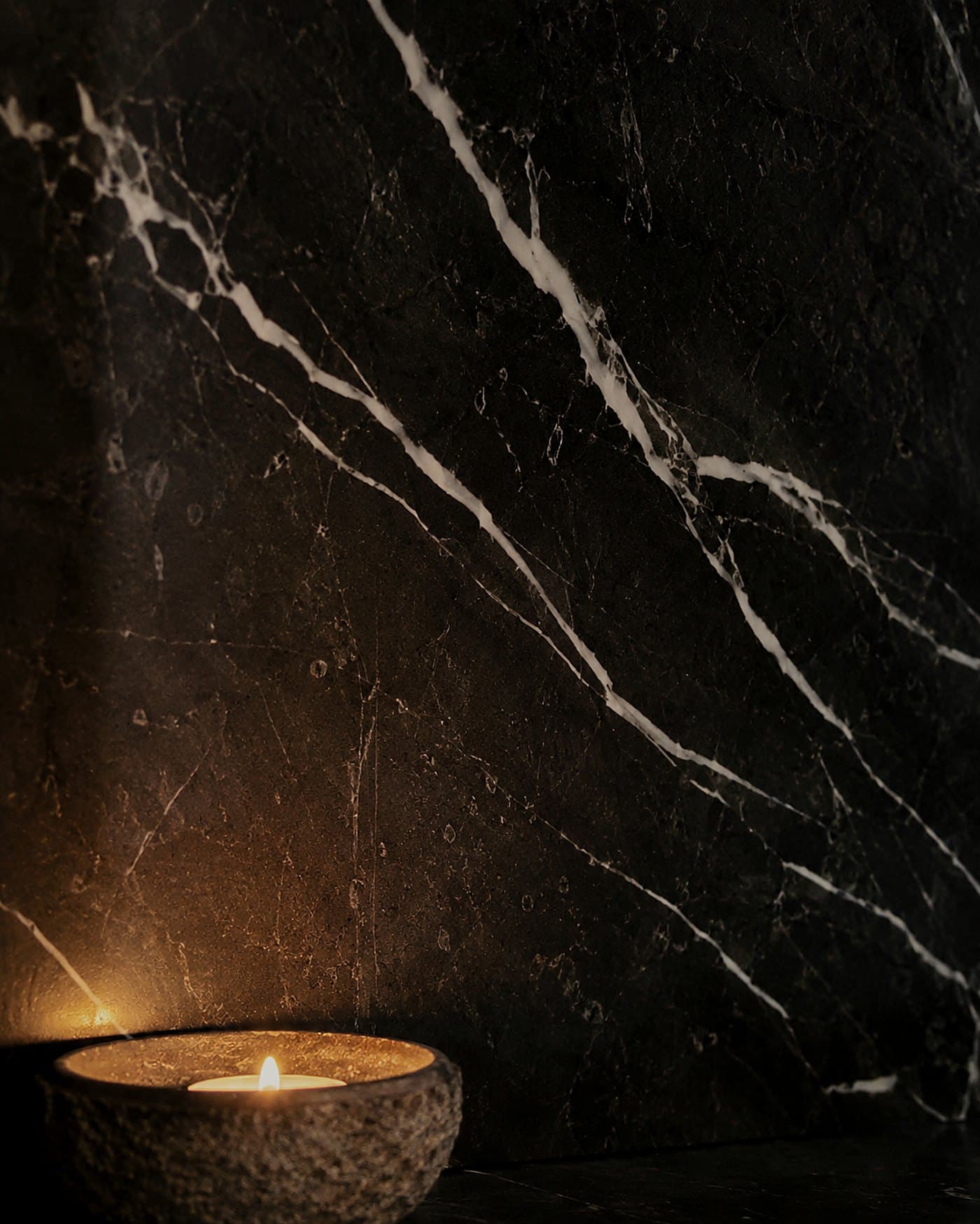Honed Nero Marquina marble with diagonal white veining in candlelight, a candle in a rough stone dish below — material study by LKYOOB