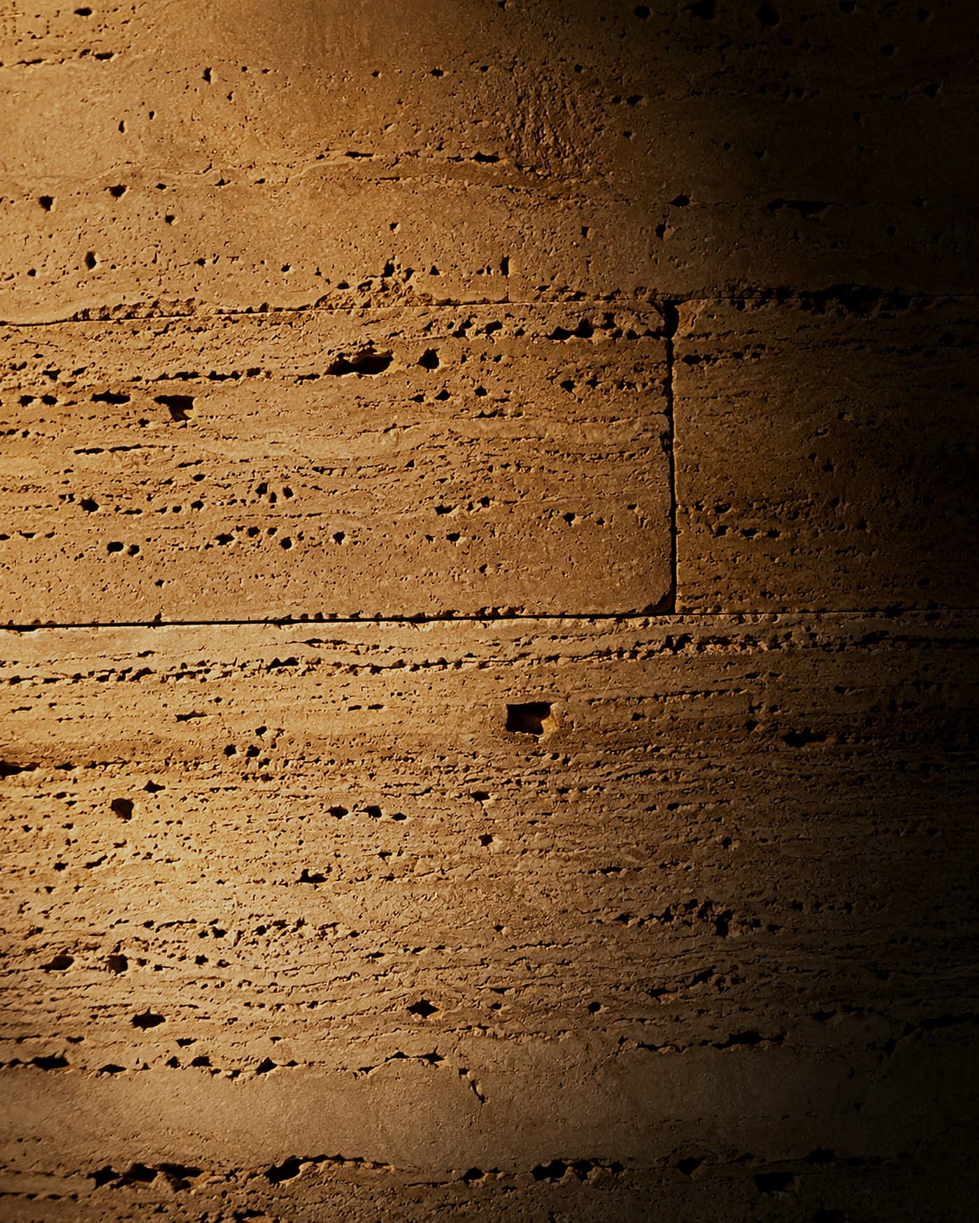 Roman travertine wall under strong warm raking light, every pore carved into relief — material study by LKYOOB